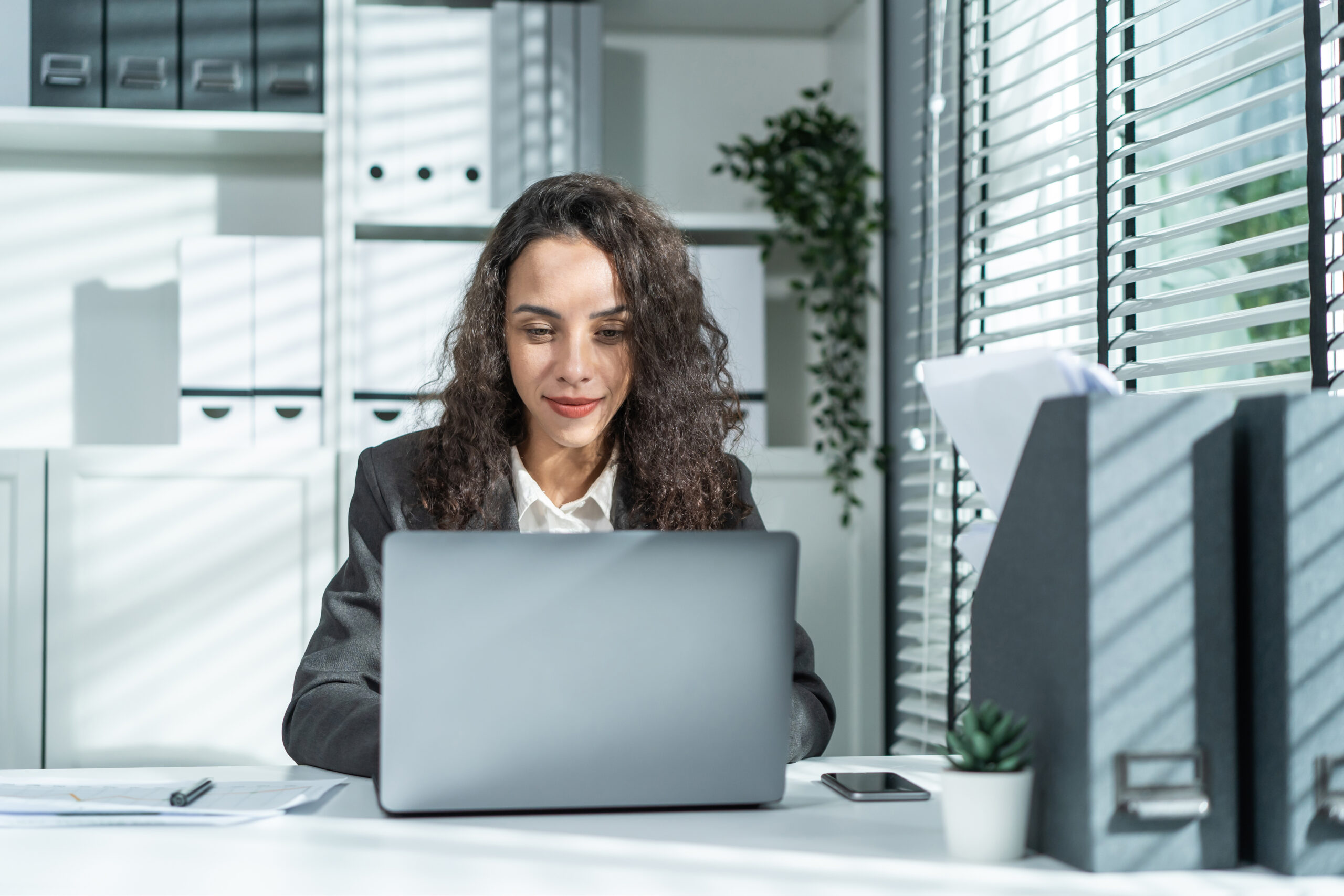 Portrait of Latino beautiful business woman smile while work in office. Attractive professional female employee worker in formal wear sit on table at workplace, use laptop computer and look at camera.